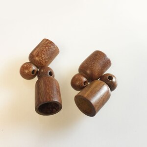 Puede incluir: Una colecci&oacute;n de peque&ntilde;as cuentas y componentes cil&iacute;ndricos de madera hechos a mano. Los componentes son de un rico color marr&oacute;n c&aacute;lido, con vetas de madera visibles. Las cuentas tienen una forma suave y redondeada, y los cilindros tienen una base acampanada y un agujero.