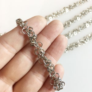 May include: Close-up of a silver-colored chain with small, interconnected circular links. The chain is held in a hand, with additional chains visible in the background. The links are approximately 0.25 inches in diameter. The jewelry is on a white background.