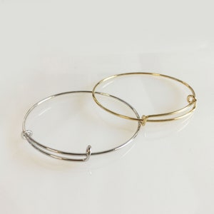 May include: Two expandable bangle bracelets. One is silver-colored, and the other is gold-colored. The bracelets are made of thin metal and have a simple, minimalist design. They are shown on a white surface.