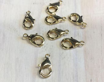 Puede incluir: Una colección de cierres de langosta dorados, ideales para la fabricación de joyas. Los cierres están dispuestos sobre una superficie de madera de color claro. Cada cierre tiene un mecanismo de resorte para un cierre seguro y un pequeño lazo.