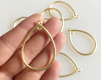 Puede incluir: Componentes de joyería dorados en forma de lágrima. Los colgantes con la parte trasera abierta tienen un lazo en la parte superior para sujetarlos a otros componentes de joyería. Varias piezas son visibles, una de ellas sostenida en una mano.