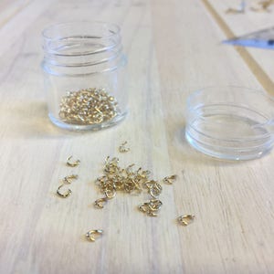 May include: A clear plastic jar filled with small, gold-colored metal clasps, with more clasps scattered on a light wooden surface. An empty plastic lid sits nearby. The clasps are likely for jewelry making or crafts.