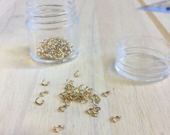 Puede incluir: Un tarro de plástico transparente lleno de pequeños cierres metálicos dorados, con más cierres esparcidos sobre una superficie de madera clara. Una tapa de plástico vacía está cerca. Los cierres son probablemente para la fabricación de joyas o manualidades.