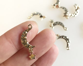 May include: A gold-coloured crescent moon charm with a textured surface and a dangling star. The charm has a small loop at the top for attaching to jewellery. Other similar charms are scattered in the background.