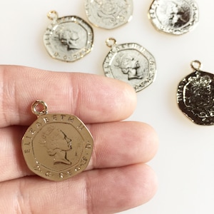 Puede incluir: Colgantes de monedas doradas con un lazo para hacer joyas. Los colgantes presentan un perfil de una persona y el texto "ELIZABETH II D.G. REG. F.D." en la moneda. Las monedas tienen un borde texturizado y están sobre una superficie blanca.