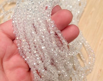 May include: Strands of clear, faceted beads are held in a hand, showcasing their sparkling, iridescent quality. The beads are strung together, creating a delicate and shimmering effect. The background is a light wooden surface.