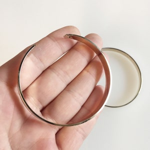 Könnte beinhalten: Ein goldfarbenes, offenes Armreif-Armband in einer Hand, mit einem zweiten Armband im Hintergrund. Die Armbänder haben ein schlichtes, minimalistisches Design und scheinen aus Metall zu sein.