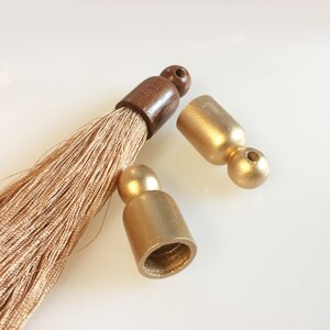 Puede incluir: Primer plano de borlas decorativas con tapas de metal dorado y una parte superior de madera marr&oacute;n. Las fibras de la borla son de color beige claro. Las tapas est&aacute;n separadas de la borla, mostrando los componentes.
