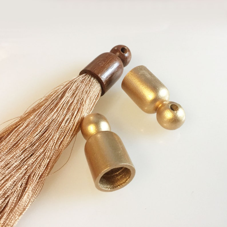 Puede incluir: Primer plano de borlas decorativas con tapas de metal dorado y una parte superior de madera marr&oacute;n. Las fibras de la borla son de color beige claro. Las tapas est&aacute;n separadas de la borla, mostrando los componentes.
