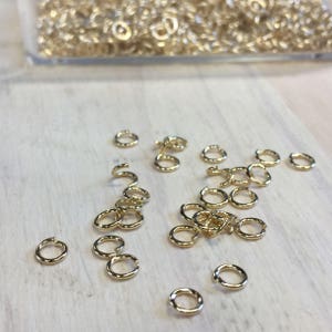 May include: A collection of small, gold-colored jump rings scattered on a light-colored wooden surface. Some rings are open, while others are closed. A clear container filled with more rings is visible in the background. These are jewelry-making supplies.