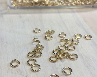Puede incluir: Una colección de pequeños anillos de salto dorados esparcidos sobre una superficie de madera clara. Algunos anillos están abiertos, mientras que otros están cerrados. Un recipiente transparente lleno de más anillos es visible en el fondo. Son suministros para hacer joyas.