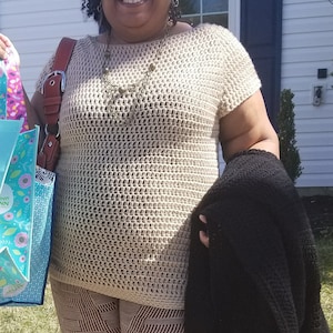 May include: A woman wearing a beige crochet top with a brown and gold necklace, striped pants, and a black cardigan. She is holding two shopping bags with floral designs.