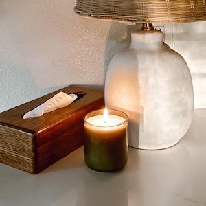 May include: A white ceramic table lamp with a woven wicker shade. The lamp is on a white countertop next to a wooden tissue box and a green candle.