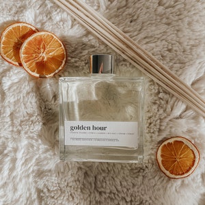 May include: A clear glass reed diffuser with a silver cap, labeled "golden hour." The label lists scents like coastal waters, citrus, and jasmine. Dried orange slices and wooden reeds are arranged around the diffuser on a fluffy, cream-colored surface.