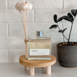 May include: A clear glass diffuser bottle with a silver cap and a label that reads "sedona" sits on a small, round wooden stand. The stand has three short legs. A bundle of reed diffuser sticks with decorative flower tops is next to the bottle.