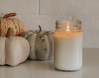 May include: A lit white candle in a clear glass mason jar with a visible flame. Beside the candle are three decorative pumpkins: one woven from tan natural fibers, one made of beige fabric, and one grey pumpkin with a distressed finish.