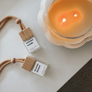 May include: Two wooden car air fresheners with white labels. The labels read "Autumn Woods" and "Favorite Flannel". Both labels include the text "homely candle co. car diffuser".
