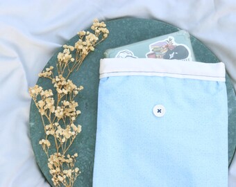 May include: A pale blue fabric pouch with a white top and a button closure. Inside the pouch is a phone with stickers. The pouch is on a dark green circular surface, with dried flowers to the left.