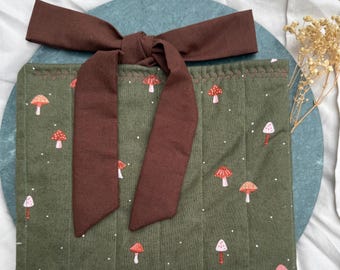 Op de afbeelding: Een groene, gewatteerde stoffen tas met een bruin lint eromheen gebonden. De tas is versierd met een patroon van witte stippen en rode en witte paddenstoelen.