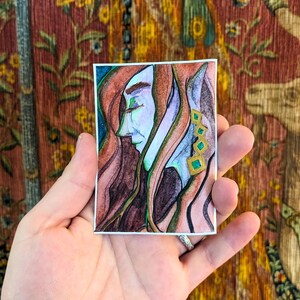 May include: A small watercolour painting depicting a person's profile with long red hair and a pale blue face. The artwork includes green, gold, and purple accents. The painting is held in a hand against a tapestry background.