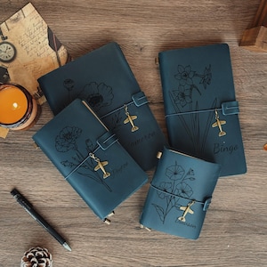 Könnte beinhalten: Vier teal-farbene Lederjournale mit floralen Designs und Flugzeug-Anhängern auf einer Holzoberfläche. Jedes Journal hat eine andere Blumenillustration und einen Namen. Eine brennende Kerze und ein Stift sind ebenfalls sichtbar.