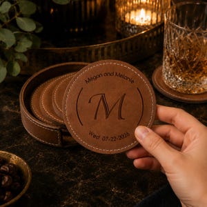 May include: A round brown leather coaster with white stitching around the edge. The coaster is engraved with the letter M in the centre, the text Megan and Melanie above, and the date Wed 07-22-2026 below. A matching holder contains more coasters.