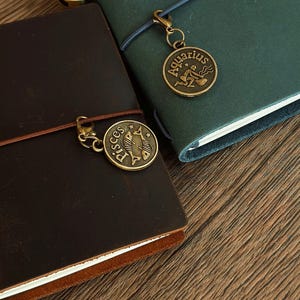 Może przedstawiać: Dwa brązowe sk&oacute;rzane dzienniki z mosiężnymi wisiorkami zodiaku. Dziennik po lewej stronie ma wisiorkiem Ryb, a dziennik po prawej stronie ma wisiorkiem Wodnika.