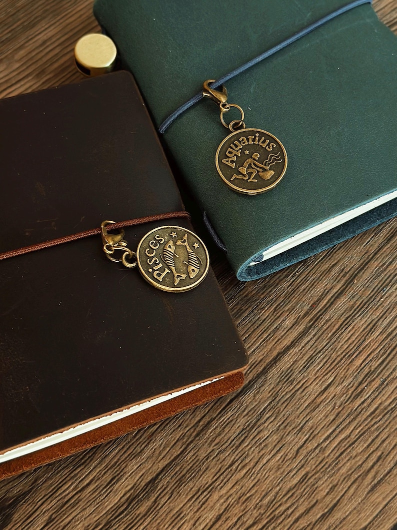 Może przedstawiać: Dwa brązowe sk&oacute;rzane dzienniki z mosiężnymi wisiorkami zodiaku. Dziennik po lewej stronie ma wisiorkiem Ryb, a dziennik po prawej stronie ma wisiorkiem Wodnika.