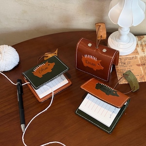 May include: Leather memo pad holders in brown and green with a pig design and names. One holder is a box shape. The memo pads have lined paper. A ball of white yarn and a black pen are also on the wooden surface.