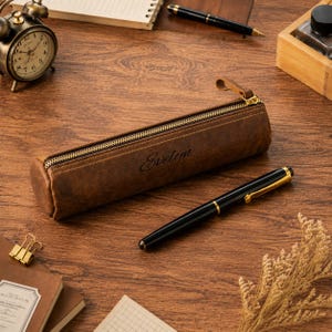 Peut inclure: Une trousse à crayons cylindrique en cuir marron avec une fermeture éclair dorée et le nom Evelyn gravé en écriture noire sur le côté. L'étui présente une texture lisse et vieillie et est placé à côté d'un stylo plume noir et doré.