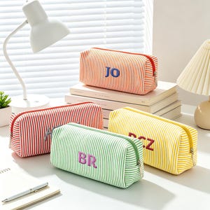 May include: Four rectangular pencil cases with a white and colored stripe pattern. Each case features a zipper closure and embroidered initials on the front. The colors include orange, red, green, and yellow, with initials reading JO, H, BR, and BCZ.