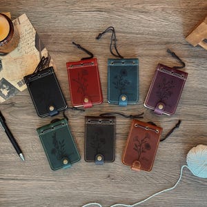 May include: Seven rectangular leather card holders in black, red, teal, purple, green, brown, and tan. Each holder features a floral line art engraving, a metal snap button closure, a top drawstring cord, and visible stitching along the edges.