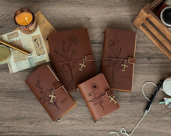 May include: A collection of brown leather journals with floral designs and airplane charms. The journals are arranged on a wooden surface with a lit candle, pen, and other decorative items. Each journal has a different floral design.
