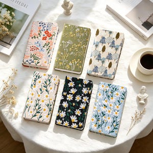 May include: Six rectangular notebooks featuring embroidered fabric covers. Designs include a pink floral pattern, a green botanical print, a blue and white tree motif, and three variations of white daisy patterns on beige, black, and light blue backgrounds.