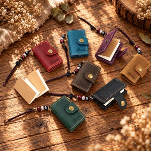 May include: A collection of small, rectangular leather notebooks in red, blue, purple, brown, tan, black, and green. Each notebook features a gold-tone snap button closure, a beaded cord strap, and a personalized name engraved on the front cover.