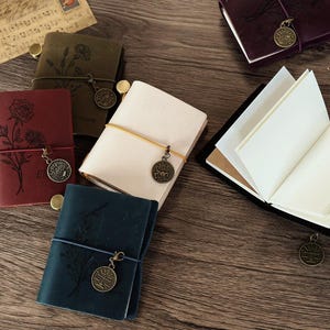Może przedstawiać: Pięć r&oacute;żnych sk&oacute;rzanych notes&oacute;w z ozdobnymi wzorami kwiatowymi i metalowymi zawieszkami. Notesy są w r&oacute;żnych kolorach, w tym brązowym, czerwonym, niebieskim i białym.