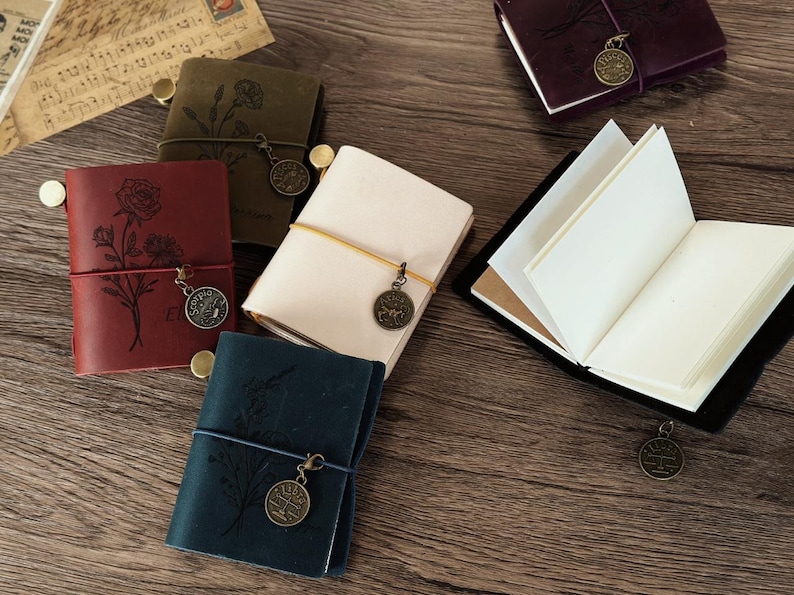 Może przedstawiać: Pięć r&oacute;żnych sk&oacute;rzanych notes&oacute;w z ozdobnymi wzorami kwiatowymi i metalowymi zawieszkami. Notesy są w r&oacute;żnych kolorach, w tym brązowym, czerwonym, niebieskim i białym.