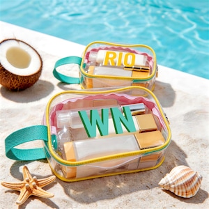 Op de afbeelding: Twee transparante cosmetische tassen met gele randen en turkooizen handvatten, gevuld met schoonheidsproducten. Eén tas heeft "RIO" in gele letters, de andere "WIN" in turquoise. Een zeester, schelp en halve kokosnoot liggen in de buurt.