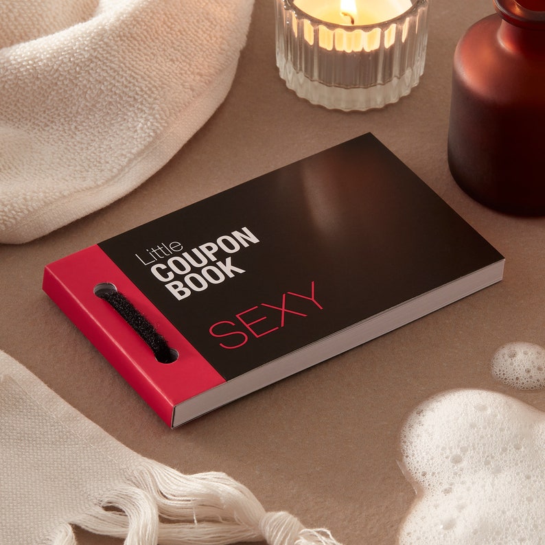 May include: A black and red "Little Coupon Book" with the word "SEXY" in red. The book has a black loop closure. A lit candle, a brown bottle, and a white towel are also in the image.
