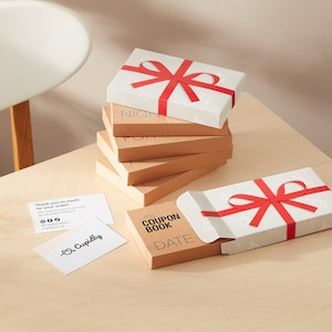May include: A collection of gift boxes tied with red ribbon. The top box is white with a pattern, the others are brown. One box is open, displaying a "Little Coupon Book" with the word "DATE". Business cards are also visible.