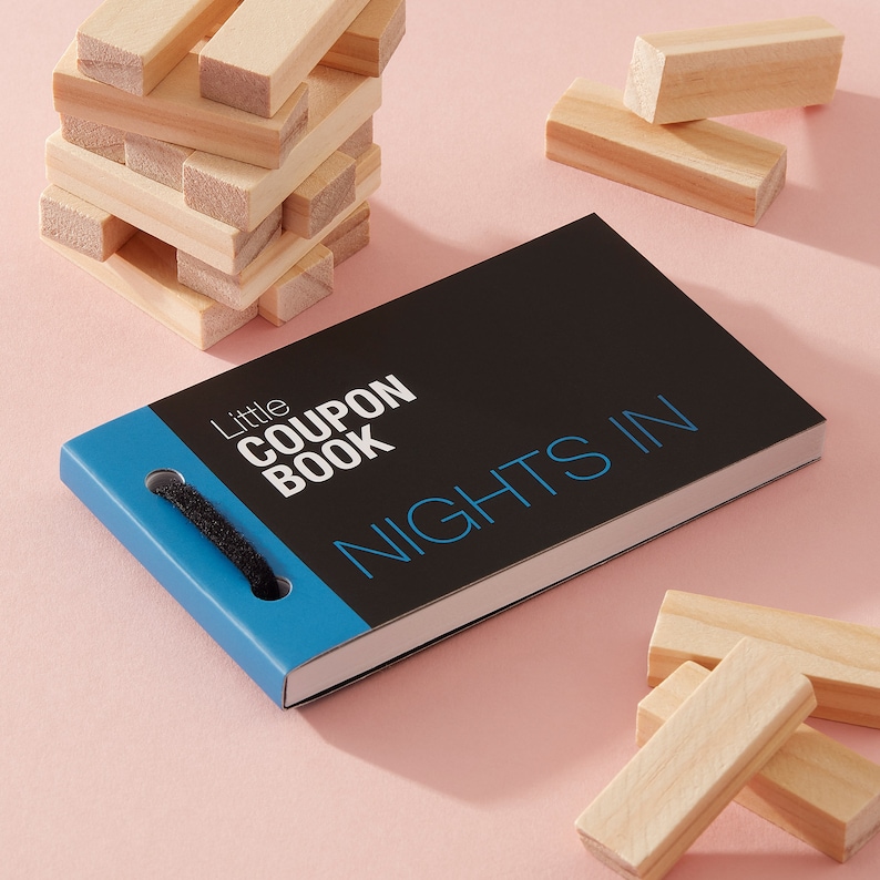 May include: A small black and blue coupon book with the text "Little Coupon Book" and "Nights In" on the cover. The book is sitting on a pink surface with wooden blocks scattered around it.