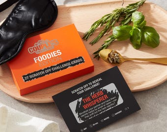 May include: A couples game called "Foodies Edition" with 27 scratch-off challenge cards. The game includes a card with the challenge "The Herb Whisperer" which instructs players to blindfold their partner and have them identify herbs by smell or taste. The card is black with white text and icons.