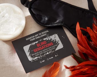 May include: A black satin sleep mask, a white bowl with ice cubes, and a black card with red text that reads "SCRATCH OFF TO REVEAL YOUR CHALLENGE! BLIND SENSATIONS Blindfold your partner and surprise them with various textures. ANY 15 min 2" along with several orange feathers.