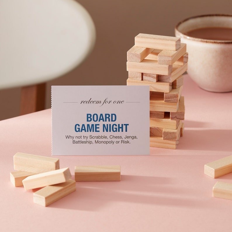 May include: A white card with the text "redeem for one BOARD GAME NIGHT Why not try Scrabble, Chess, Jenga, Battleship, Monopoly or Risk."  The card is sitting on a pink surface with a stack of wooden blocks and a cup of coffee.