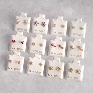 Pode incluir: Doze pares de brincos de ouro 14 quilates com diferentes desenhos de flores e folhas. Os brincos são exibidos em cartões brancos com o texto "14 KT GOLD" impresso em cada cartão.