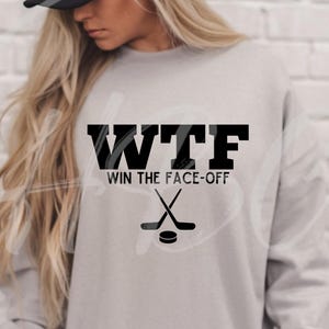 May include: A light gray long-sleeve sweatshirt featuring black text that reads "WTF WIN THE FACE-OFF" above a graphic of two crossed hockey sticks and a puck. The garment has a crew neckline and long sleeves, providing a casual look.