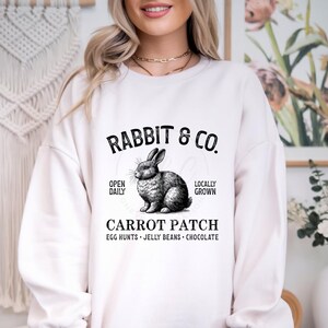 Op de afbeelding: Wit sweatshirt met een zwart-witte illustratie van een konijn. De tekst "RABBIT & CO." staat boven het konijn en de tekst "CARROT PATCH" onder het konijn. De tekst "OPEN DAILY" en "LOCALLY GROWN" staan aan weerszijden van het konijn. De tekst "EGG HUNTS - JELLY BEANS - CHOCOLATE" staat onder de tekst "CARROT PATCH."
