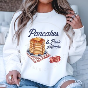 Könnte beinhalten: Weißer Pullover mit einem Grafikdruck von einem Stapel Pfannkuchen mit Sirup und Butter und dem Text "Pancakes & Panic Attacks" in Blau. Die Grafik enthält auch eine rot-weiß karierte Serviette und Blaubeeren. Unter der Grafik befindet sich ein roter Kreis mit dem Text "Buy One Get One" in Weiß.