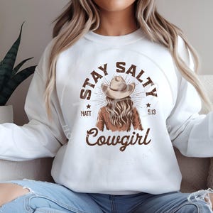 Op de afbeelding: Wit sweatshirt met een bruine grafische print van een vrouw met een cowboyhoed en de tekst "Stay Salty Cowgirl" met de woorden "Matt" en "5:13" aan weerszijden van de afbeelding.