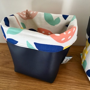 Pode incluir: Uma caixa de armazenamento de tecido azul marinho com um forro de tecido branco com um padrão abstrato colorido em rosa, azul, verde e amarelo. A caixa tem uma etiqueta branca com a palavra "Handmade" nela.
